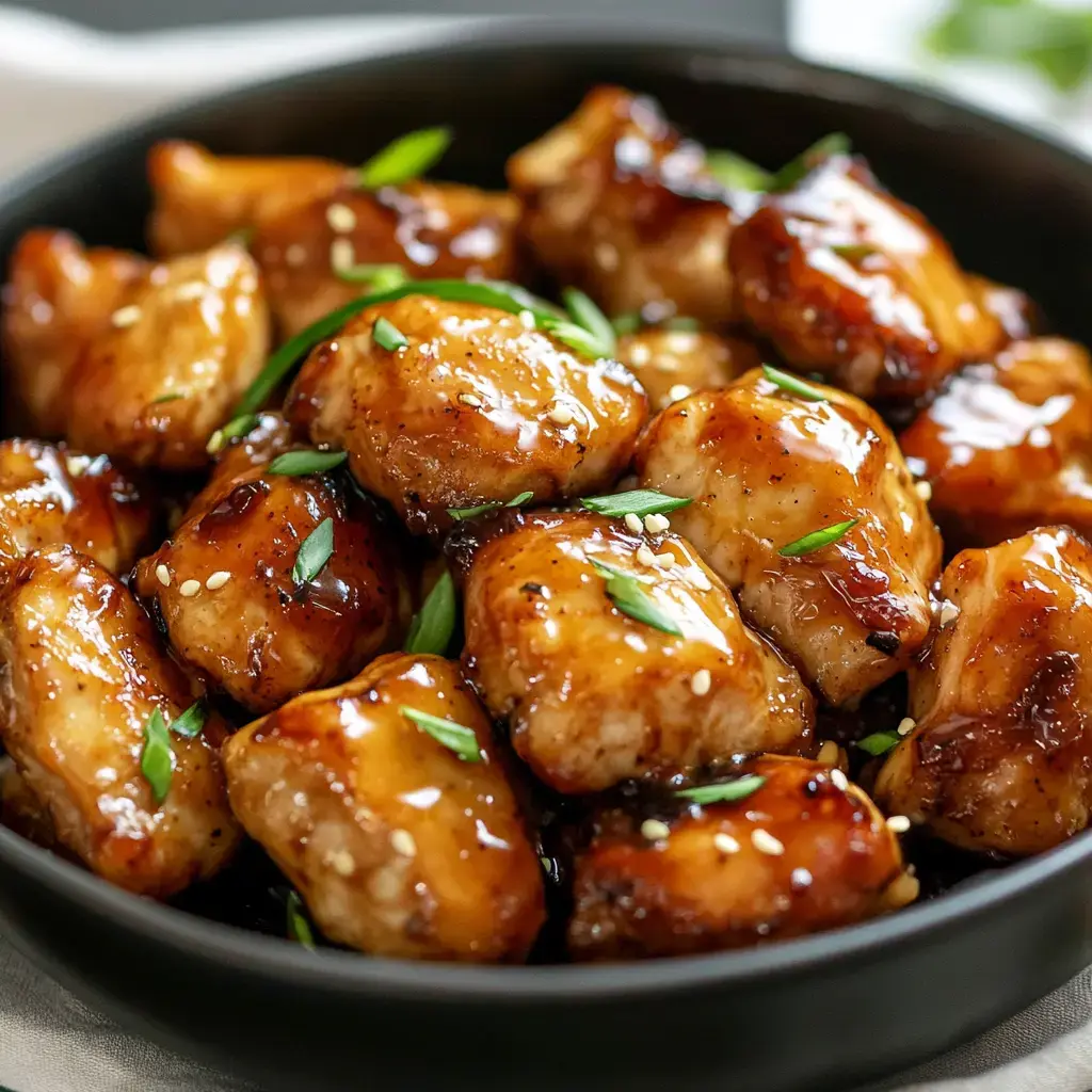 Glazed chicken chunks topped with sesame seeds and green onion served in a dark bowl.