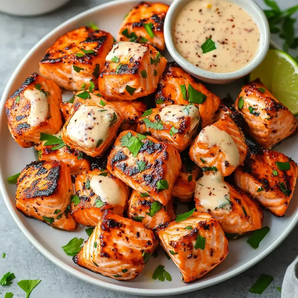 Grilled salmon cubes on a plate, garnished with parsley, with a bowl of sauce and a lime wedge on the side.