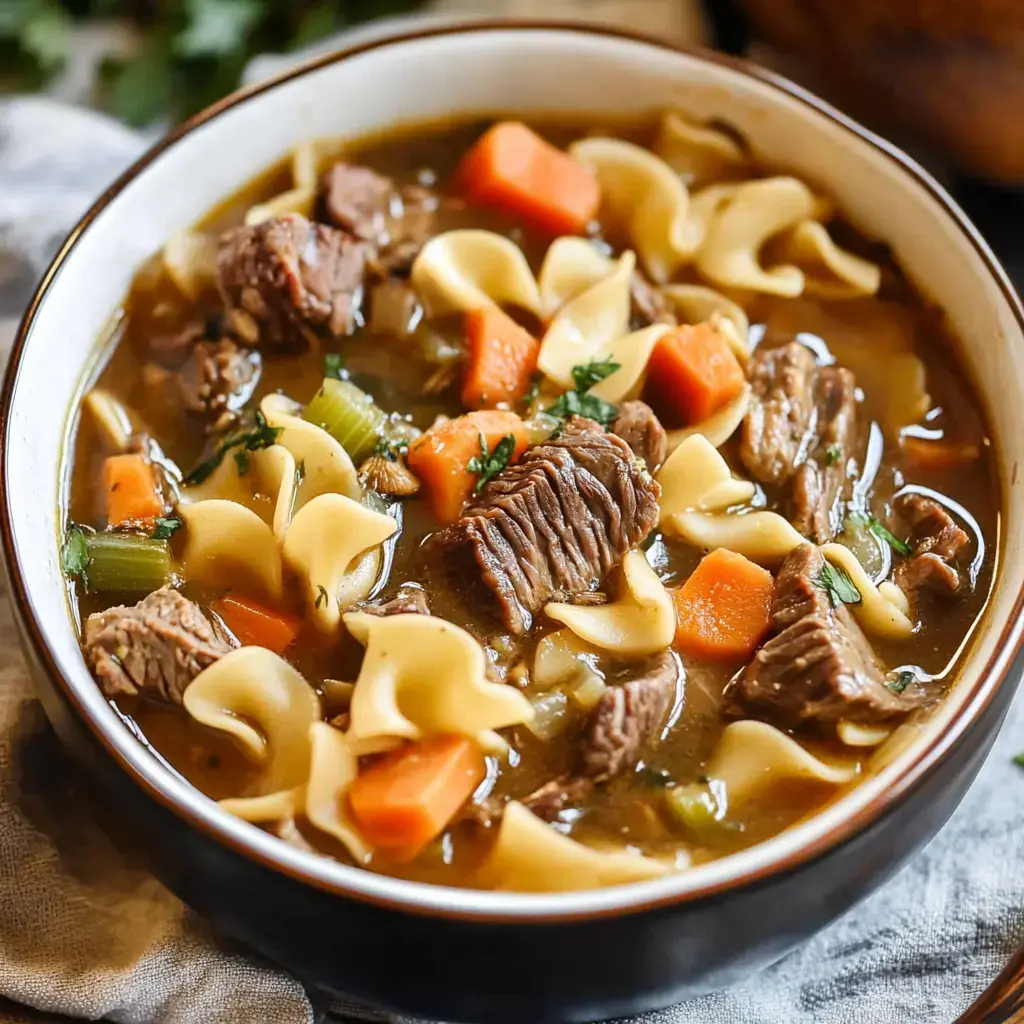 Chunks of soft beef, carrots, celery, and squiggly egg noodles floating in a steaming beefy broth, all cozied up in a big bowl.
