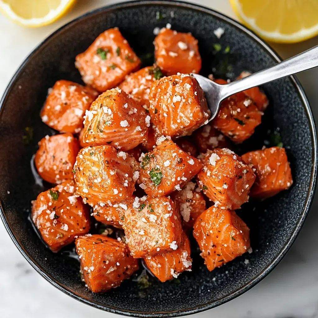 Diced seasoned salmon in a black bowl, sprinkled with herbs and salt and served with lemon wedges.
