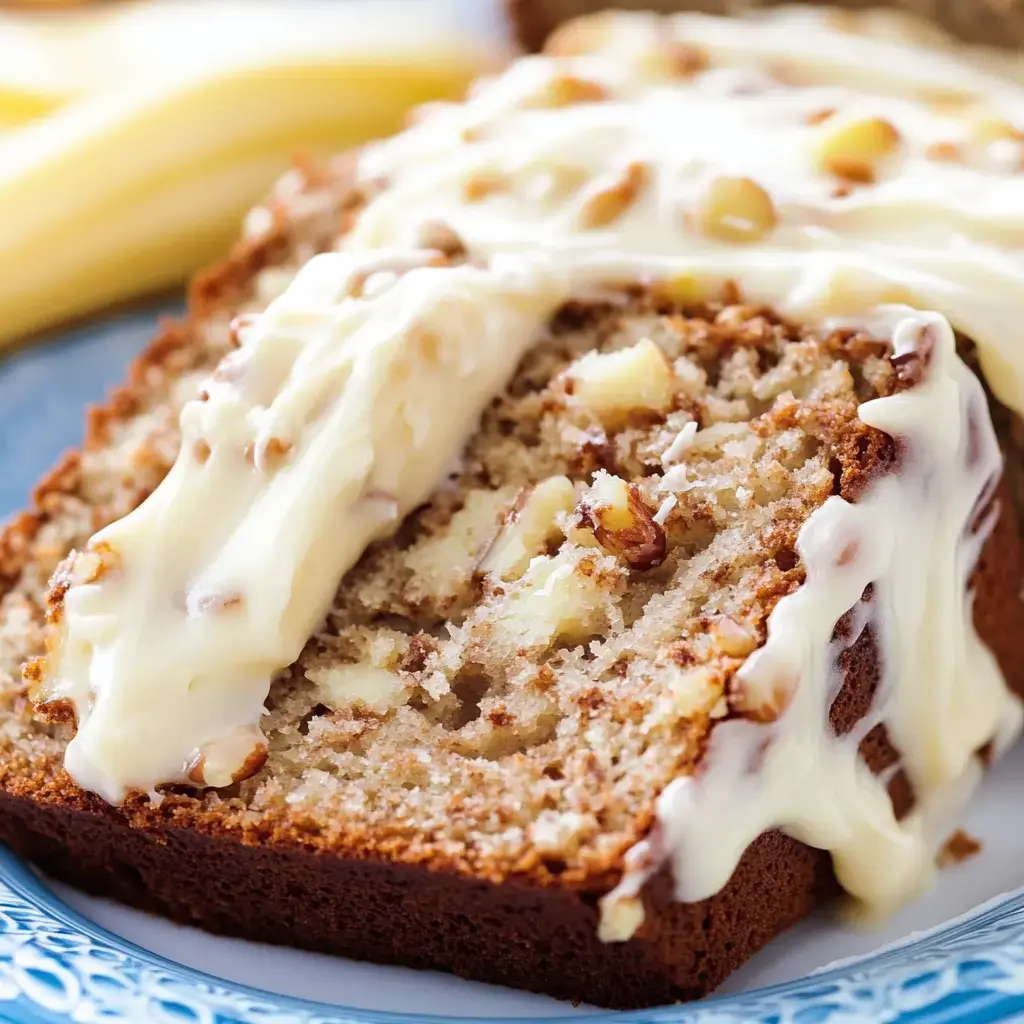 A banana bread slice covered with rich frosting and chopped nuts, presented on a blue decorative plate next to fresh bananas.