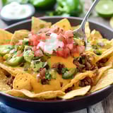 A close-up of a bowl of nachos topped with melted cheese, ground beef, jalapeños, diced tomatoes, guacamole, cilantro, and sour cream.