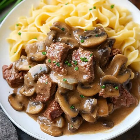 Creamy mushroom beef sitting over egg noodles, topped with green onions in a cozy serving bowl.